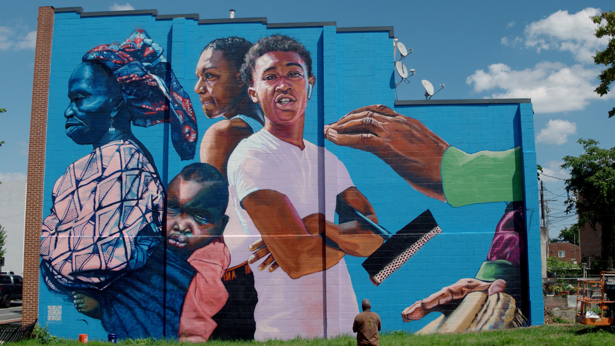 A full image showcasing the mural "African in America." On the left, a Black woman faces left in traditional clothes carries a baby on her back. In the center foreground is a young Black man in a white shirt looking at the viewer, and a Black woman behind him wearing a tank top. To the right is a hand emerging from a green sleeve playing a drum.