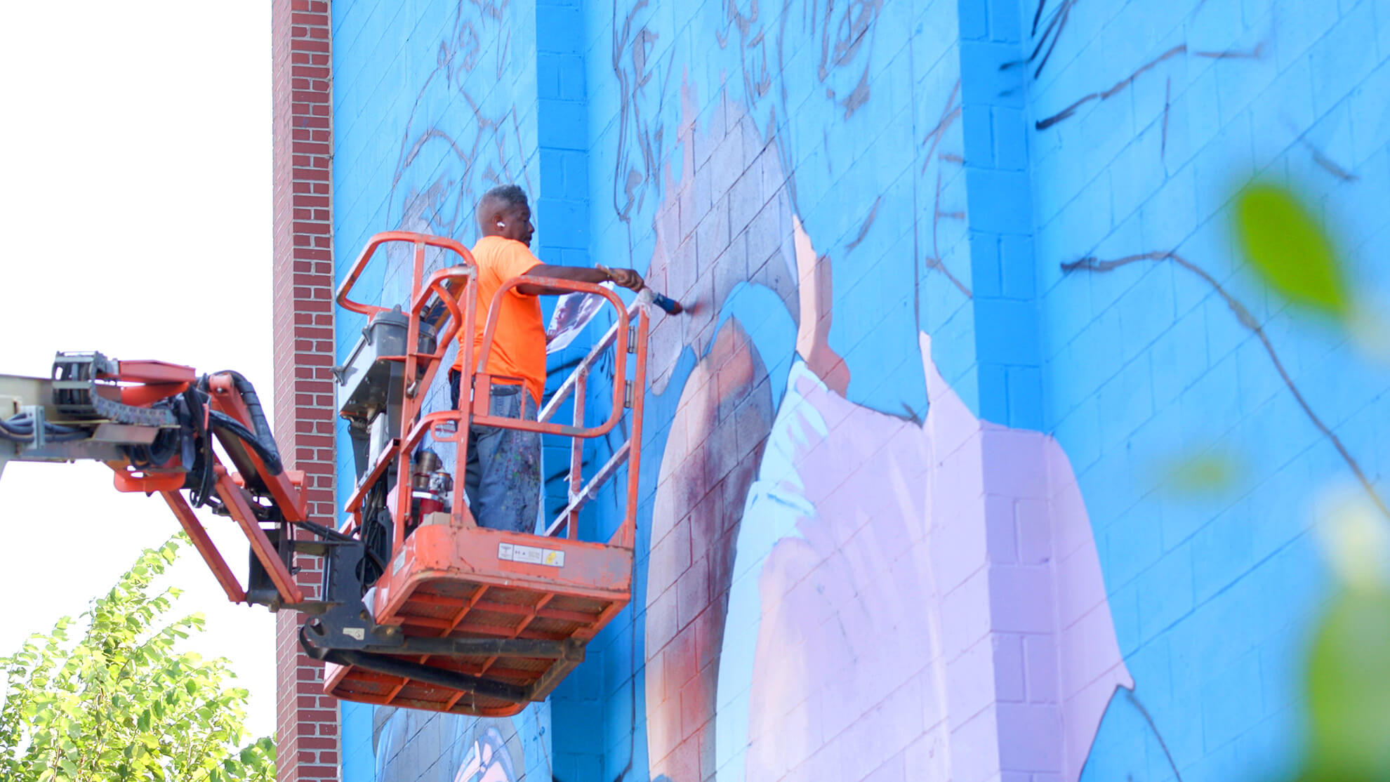 The artist Ernest Shaw, wearing an orange shirt and jeans mending the mural while on a mobile elevated working platform.