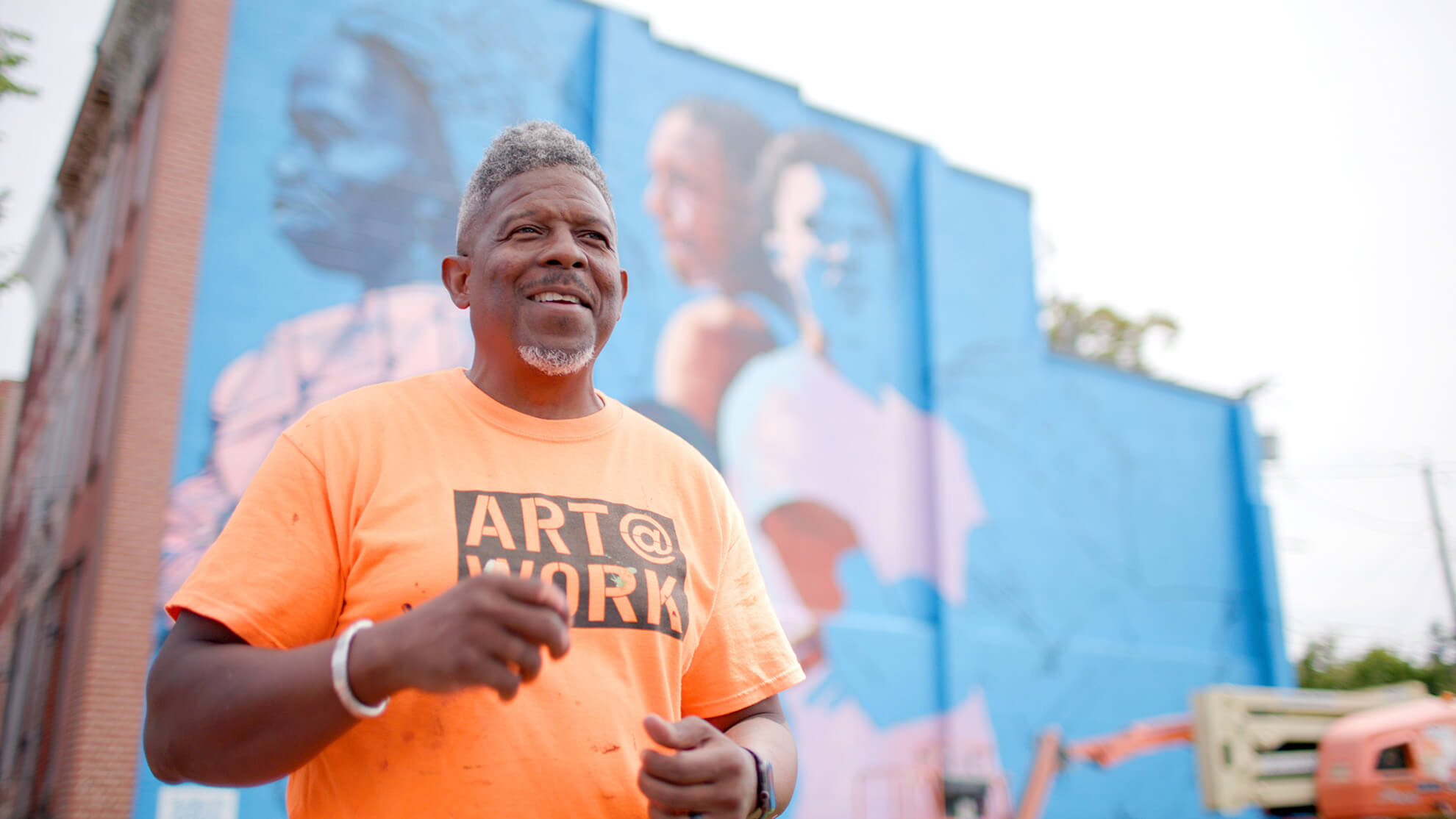 A portrait of the artist Ernest Shaw smiling and describing his work. He is a middle-aged Black man with a greying fade and neat beard. The orange shirt he wears has the phrase "Art @ Work"