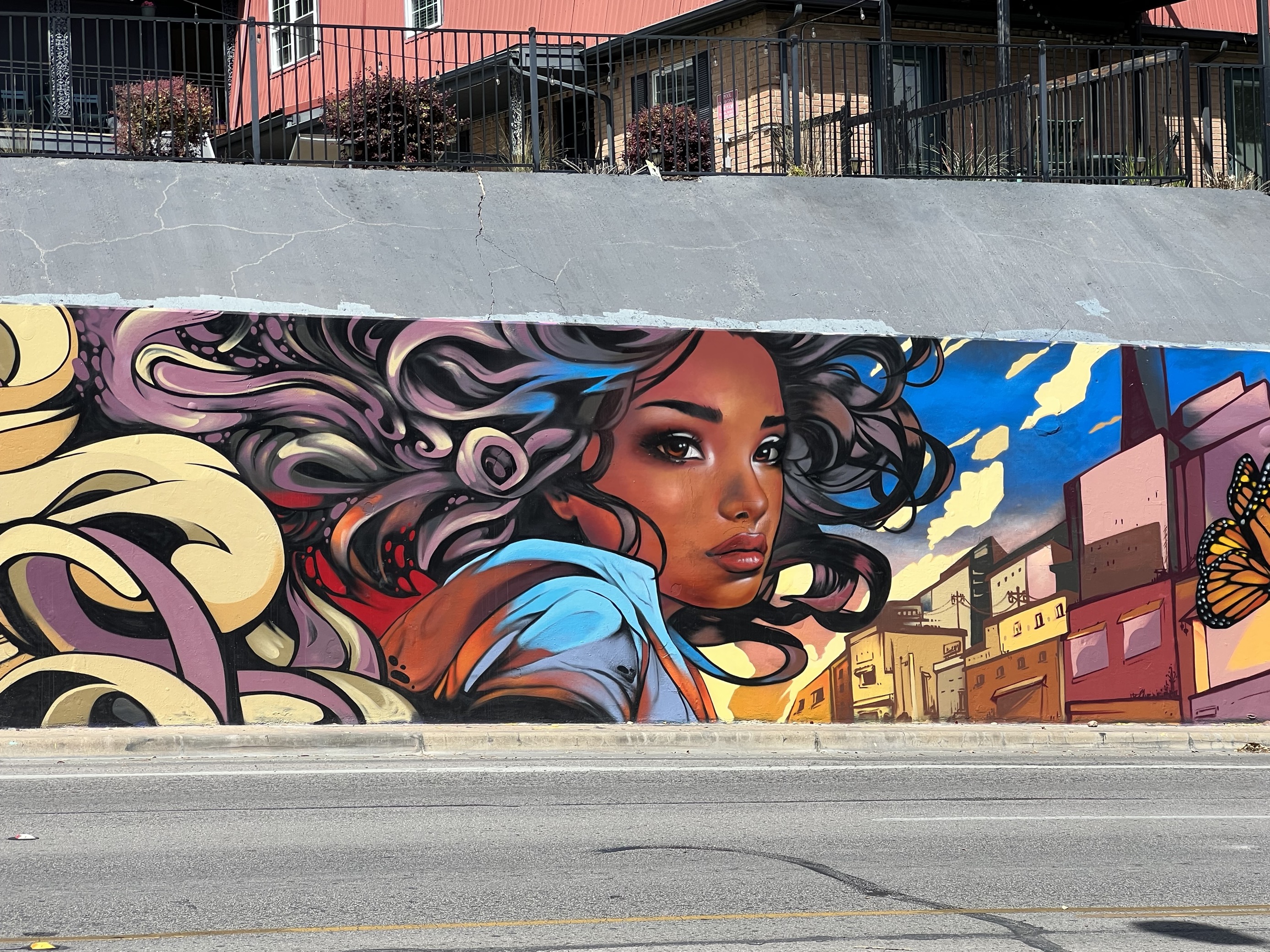 A full image showcasing the mural "Chase The Light." At the center, a Black woman faces the camera. She has long, curly hair that fades into colorful artistic designs. A monarch butterfly is depicted flying away from the woman. In the background, there are city buildings of varying color.