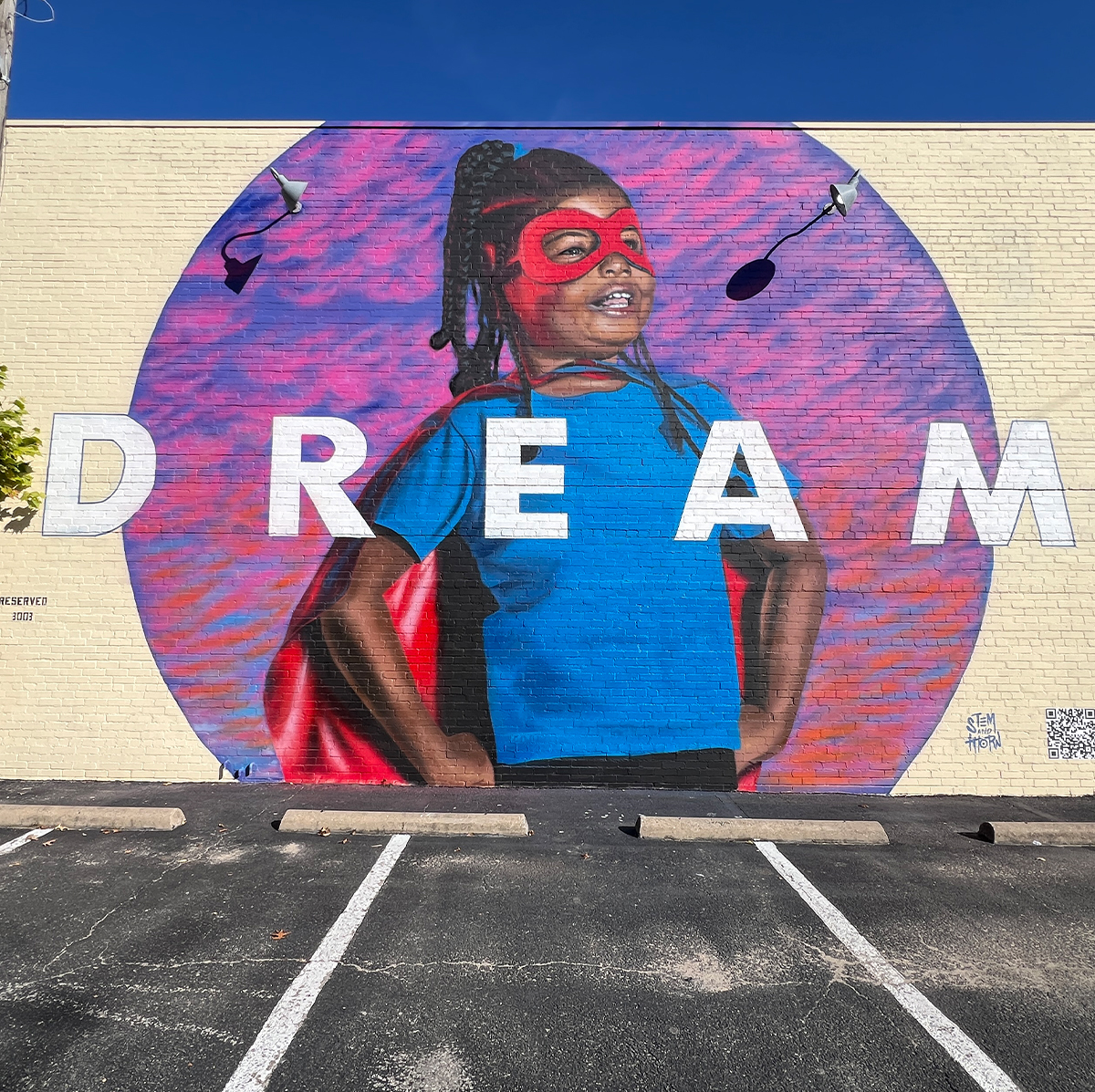 The mural "DREAM." A young Black girl is depicted with a superhero’s mask and cape, standing proudly with a big smile. Across the mural is the word "DREAM" in bold, capital letters.