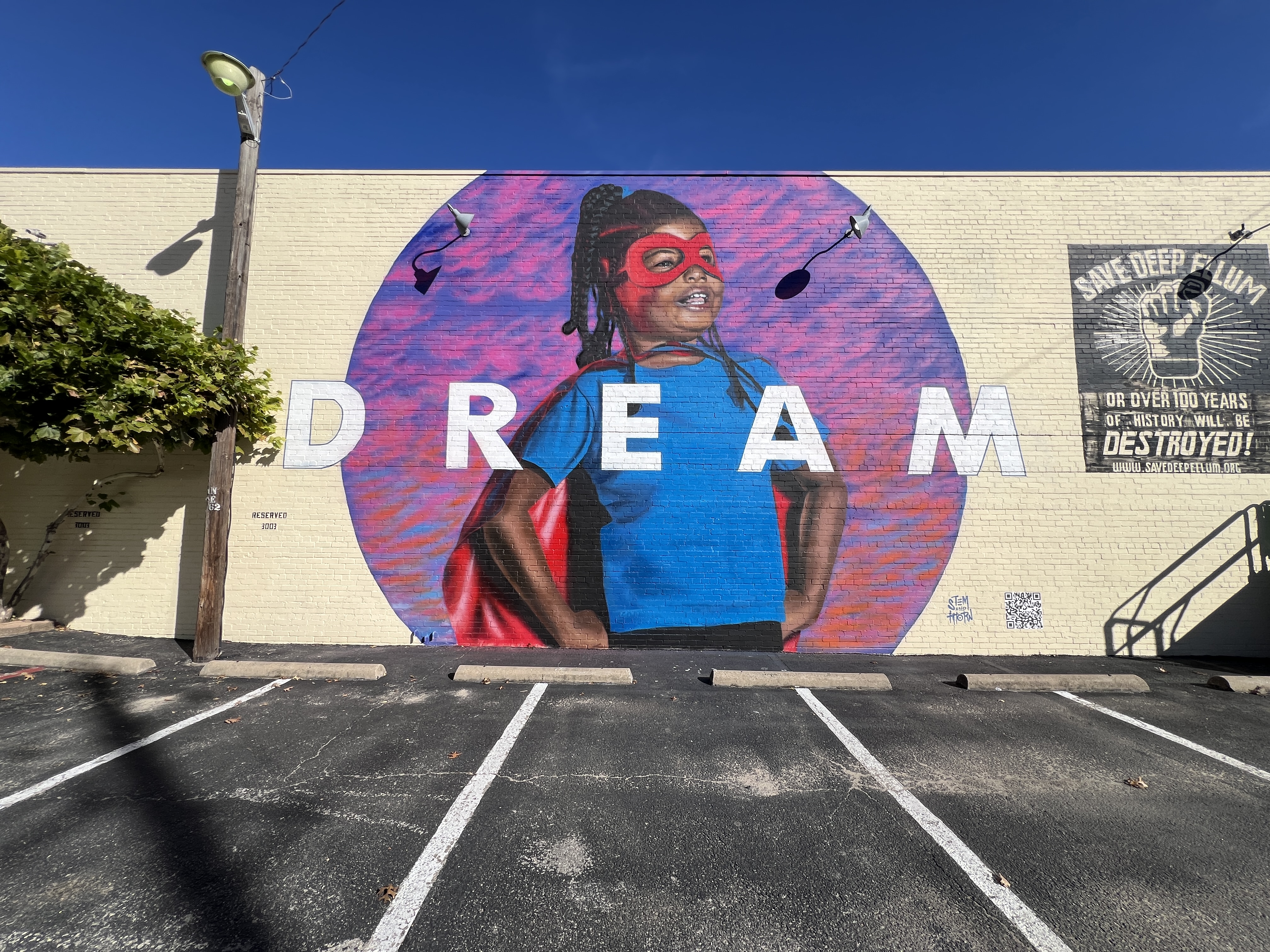 A full image showcasing the mural "DREAM." A young Black girl is depicted wearing a blue shirt and a superhero’s mask and cape. She stands proudly with her hands on her hips and has a big smile. Across the mural is the word "DREAM" in bold, capital letters.
