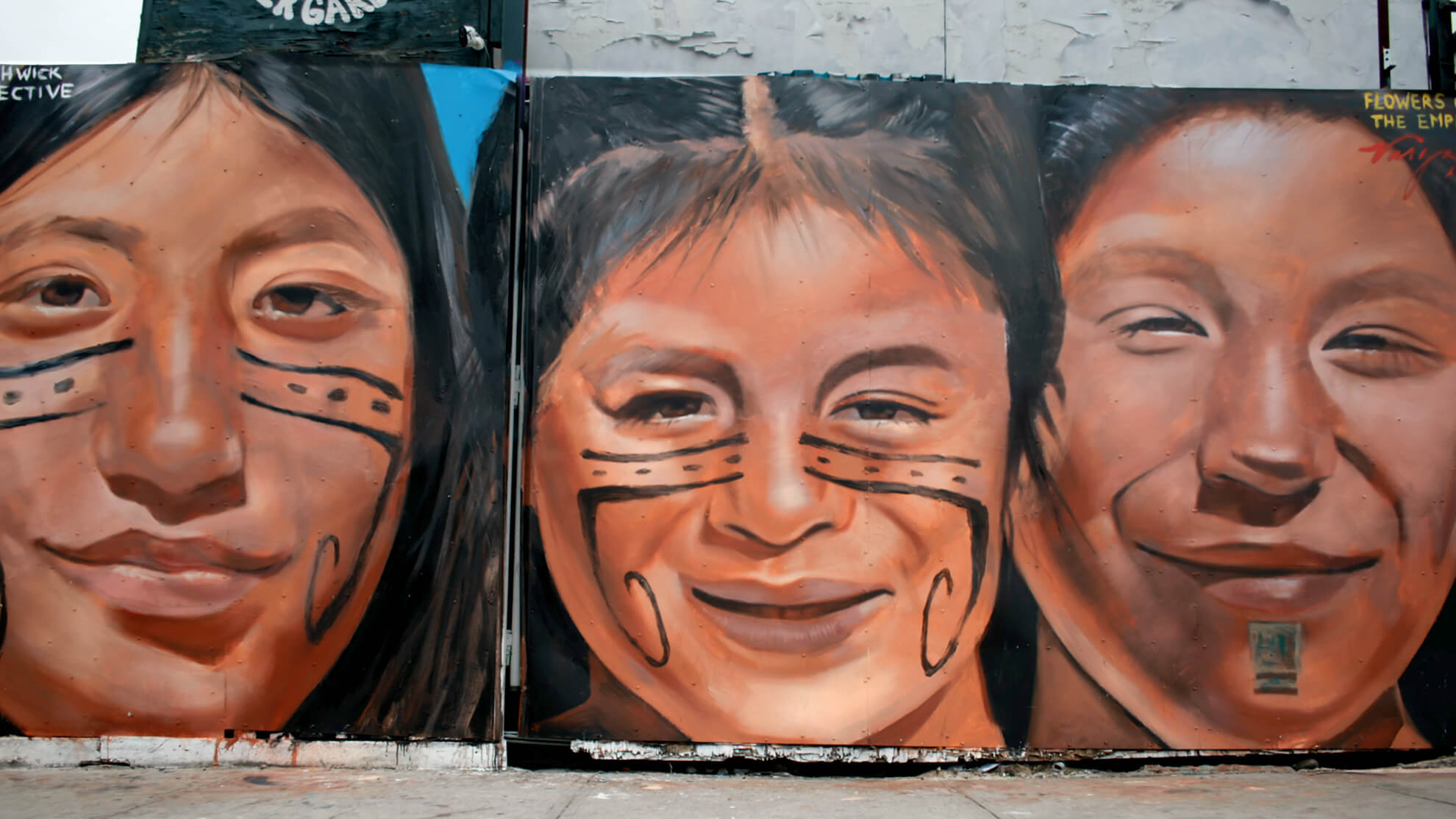A full image showcasing the mural "Flowers of the Empire." Three Indigenous faces return the viewer’s gaze. The man on the left and the woman in the center share similar facial markings, which are three black lines drawn horizontally under the eyes and starting at the bridge of the nose. The top line is solid while the middle line is dashed; the bottom line takes a sharp turn at the cheekbones and curls down next to the mouth on both sides.