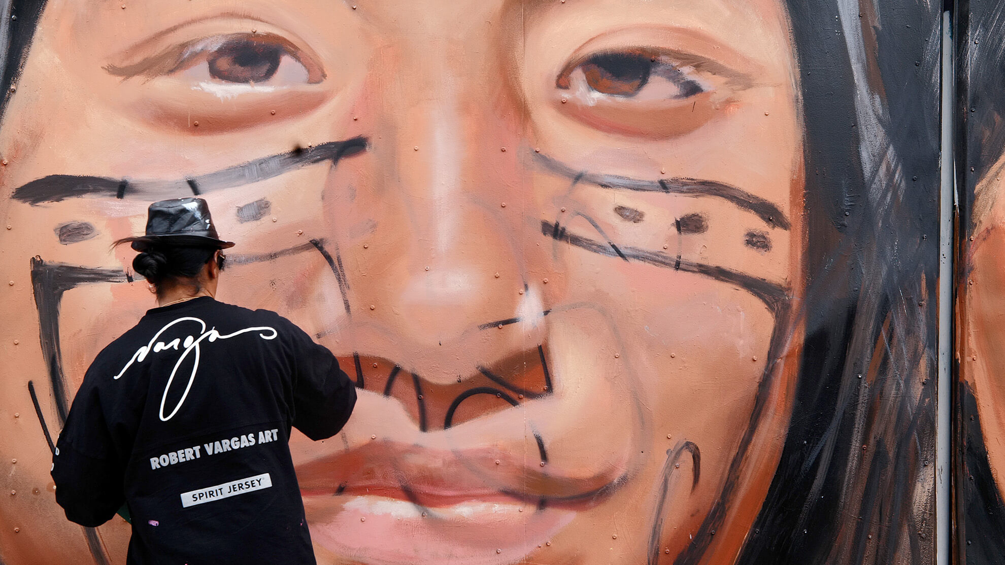 A picture of the artist from behind as he is mending the mural. He wears a hat and a black shirt bearing his name.