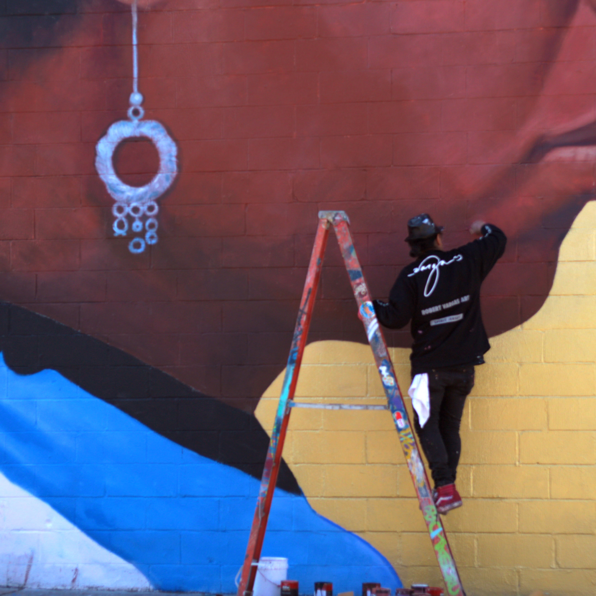 An in-progress shot of the artist mending the mural Gods That Walk Among Us. He is on a ladder and painting part of a woman's face.