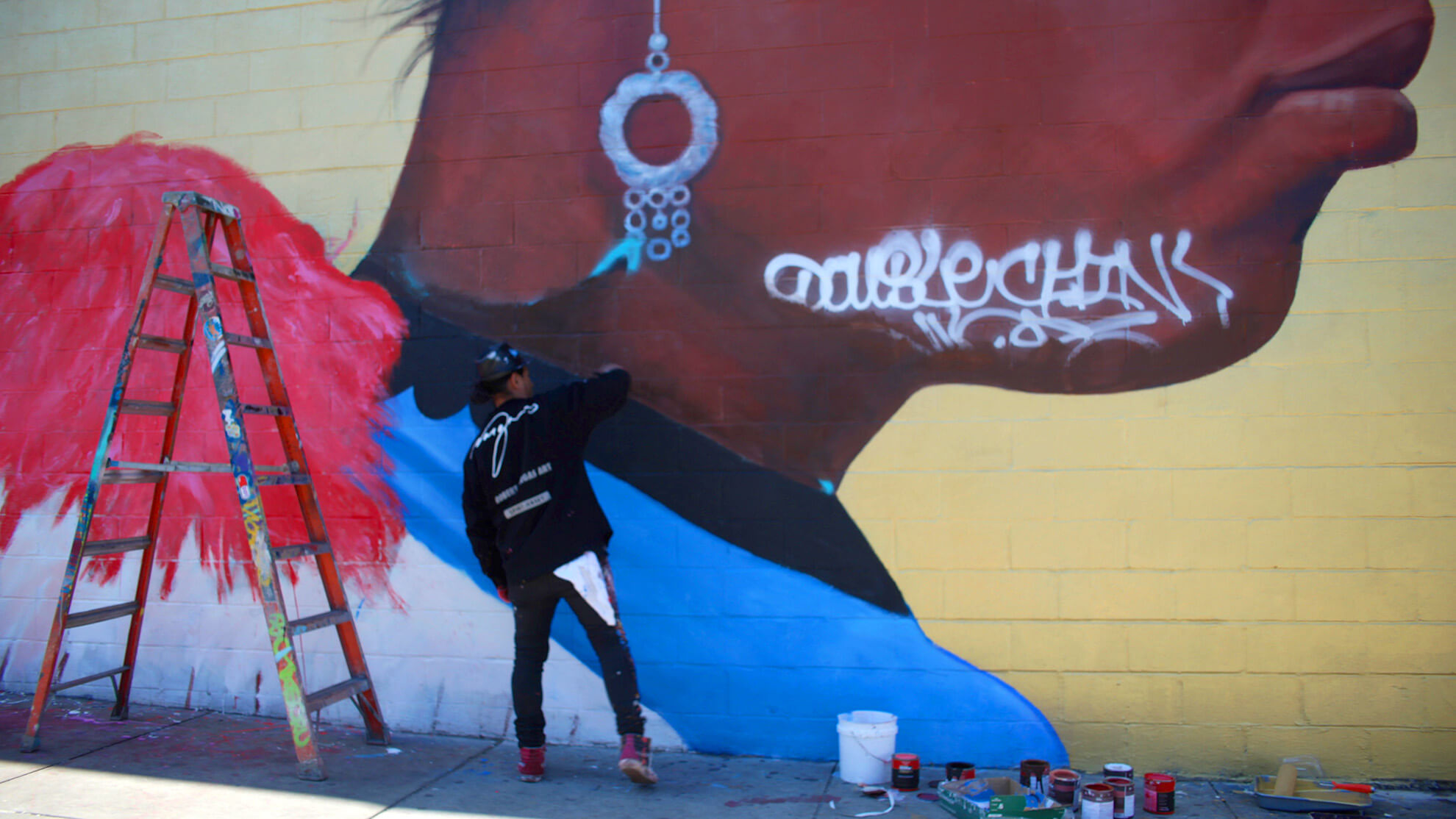 A picture of the artist, Robert Vargas, from behind has he begins to mend the mural "Gods that Walk Amongst Us." THere is graffiti on the face in the top right, obscuring it. Vargas has brought with him paints, which are to his right on the sidewalk, and to the left is a well-used ladder.