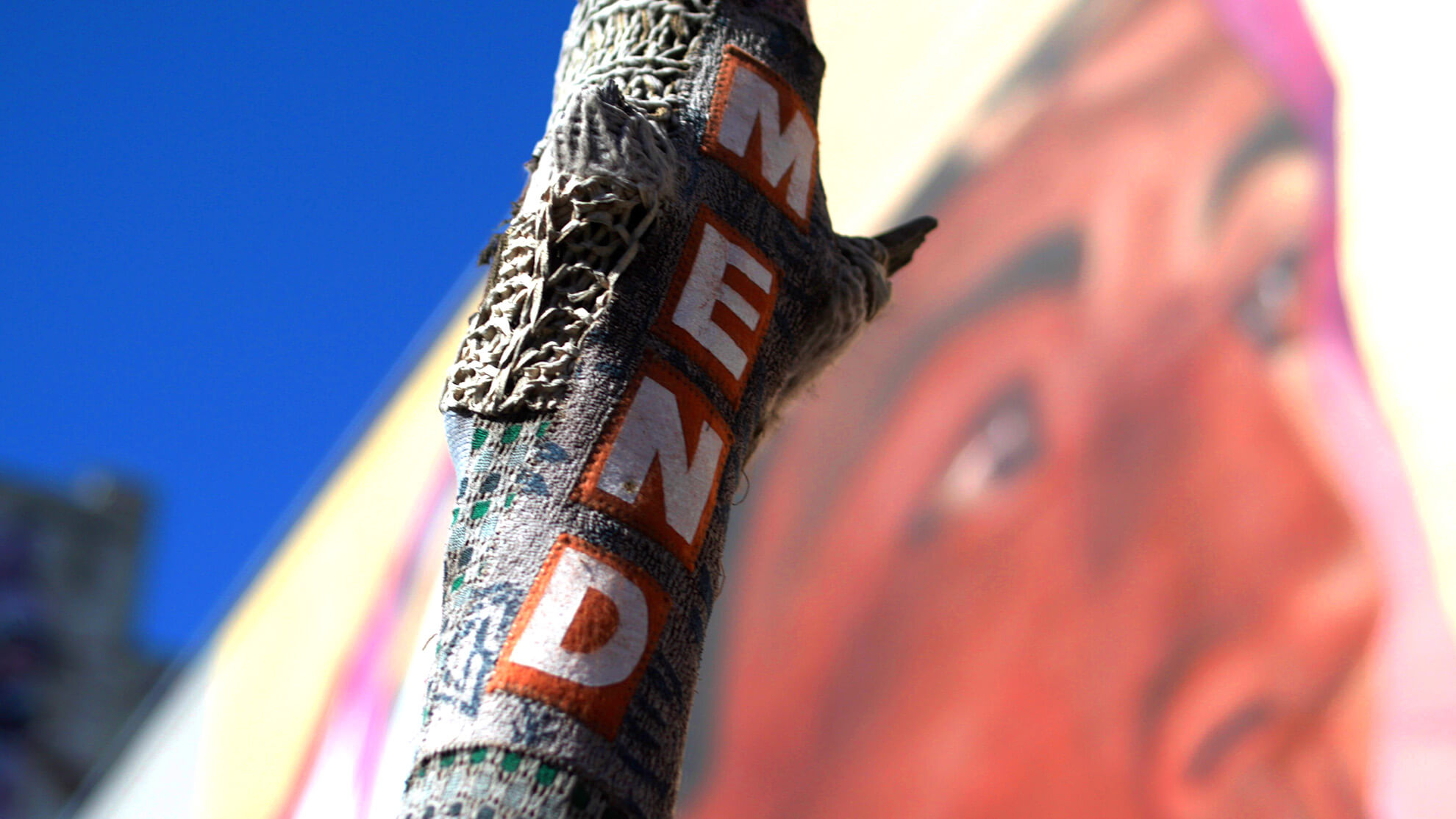 An extreme close-up of a tree with fabric art wrapped around it. The art reads "MEND", vertically, in bright white letters on an orange background. Part of the mural can be seen in the background, just out of focus.