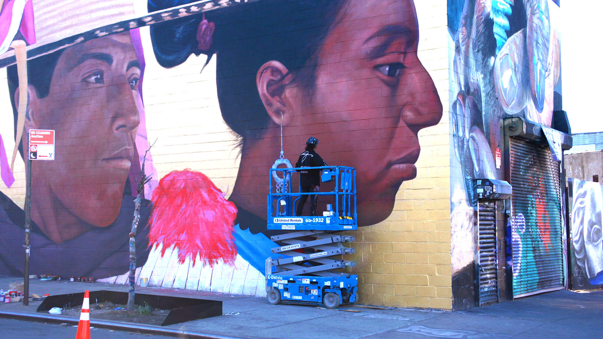 A picture of the artist from the back as he stands on a blue mobile elevated working platform that is lowering. The mural looks to be almost completely mended.