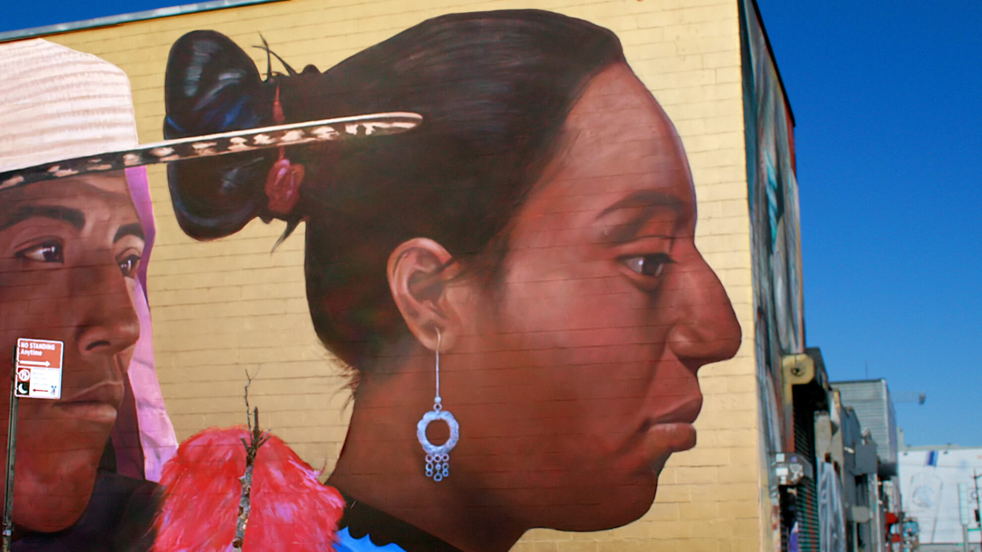A close-up of the right side of the mural, focusing on a dark-skinned Latina woman looking off to the right. Her hair is up and from her ear dangles a silver loop earring that hangs from a thin chain.