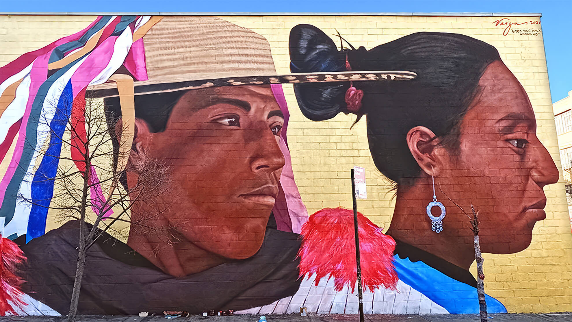 A full image showcasing the mural "Gods that Walk Amongst Us." To the left of the woman featured in the previous photo we can see a dark-skinned Latino man wearing a traditional hat also looking off to the right. His straw hat has colourful ribbons streaming from the top.