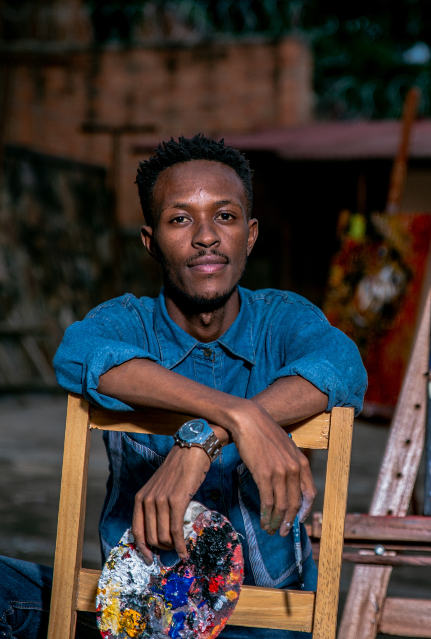 A dark-skinned Black man holding a palette and siting in a chair looking at the viewer.