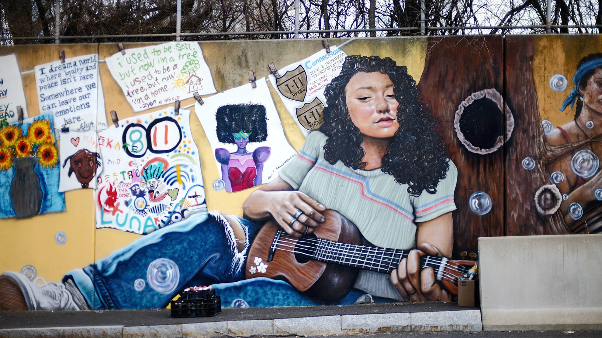 A full image showcasing part of the mural "Star Gazing." Central to the image is a light-skinned young Black woman with long, curly hair playing a ukulele while leaning against a tree trunk. To the left are numerous pictures that appear to have been drawn by children, and to the right are bubbles floating up above a Jersey barrier, which is not part of the mural.