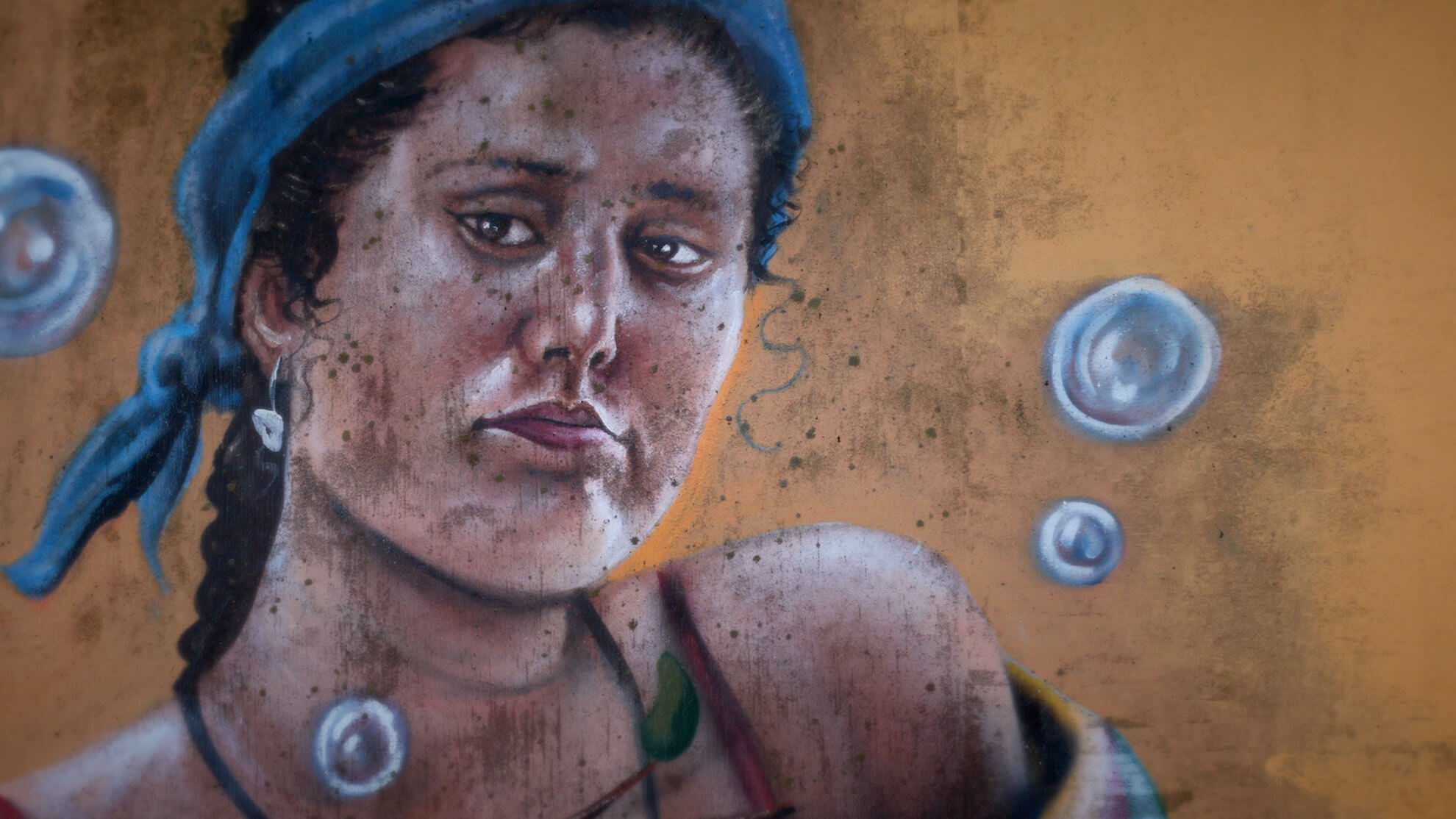 A close up of another part of the mural. A Black woman with a serious expression and wearing a blue tied headband is partially framed by bubbles on an ochre background.