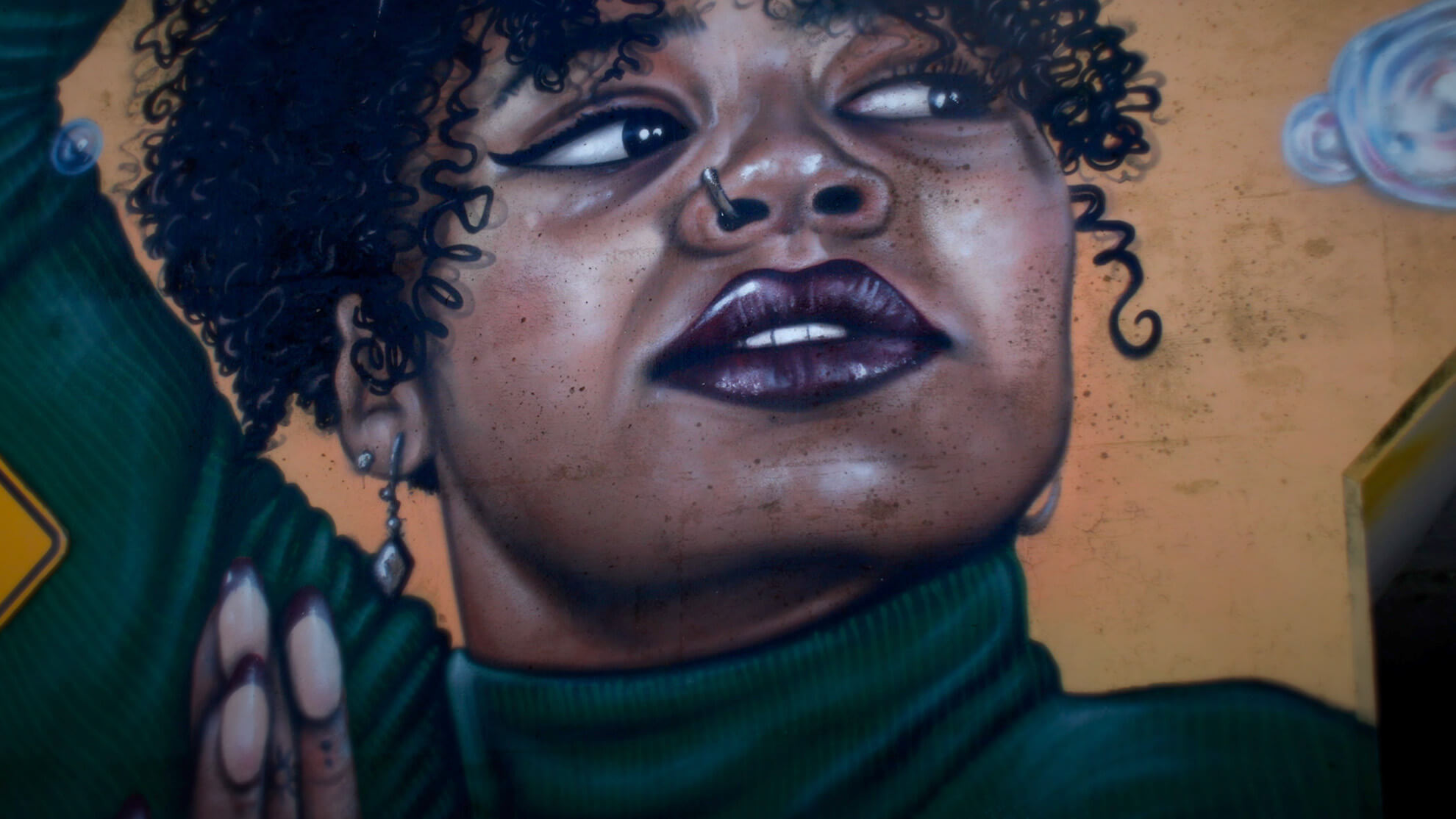 A dark-skinned Black woman with coiled hair and a nose piercing looks off into the distance in this close-up. She is wearing a deep forest green turtleneck and is also partially framed by bubbles on an ochre background.