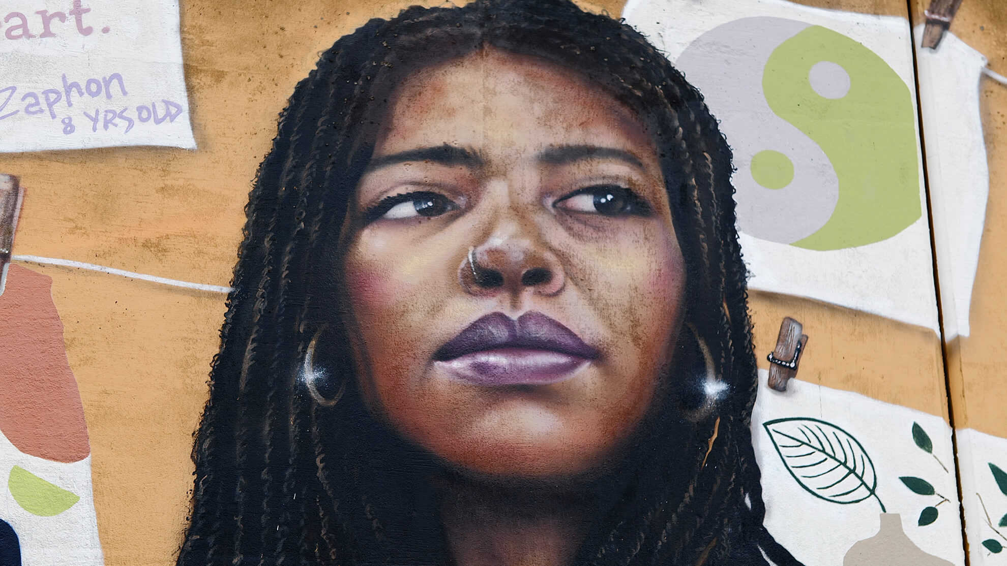 A different dark-skinned Black woman with a nose piercing and braids looks off into the distance while framed by children's pictures in the background.